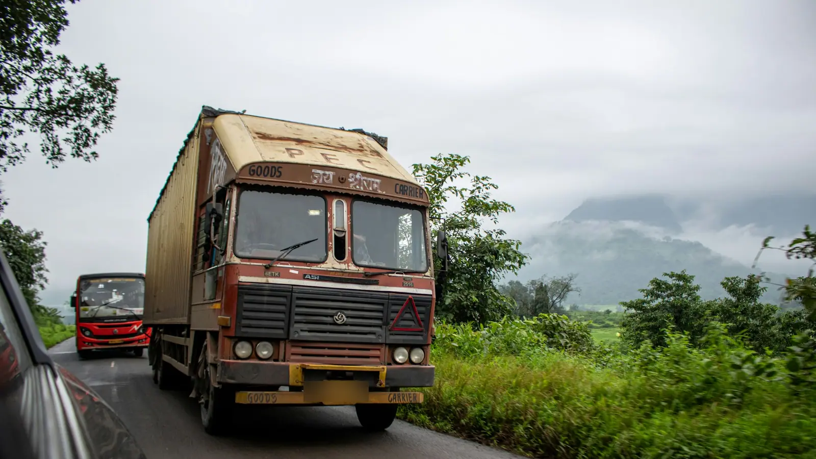 A goods truck on a wet two-lane road through green hills, with a bus behind it.