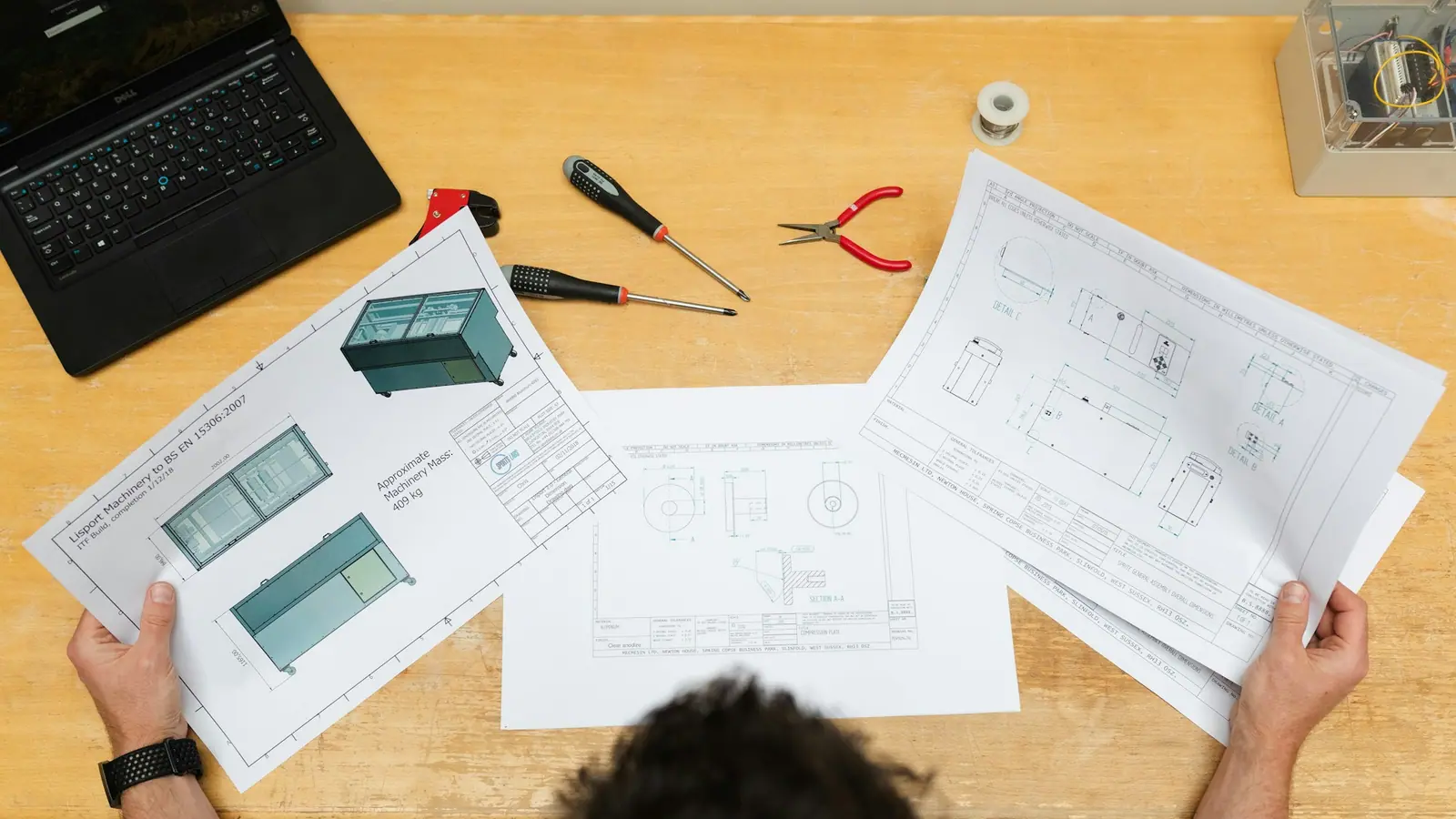 Engineers' hands holding printed CAD drawings of a part over a workbench with tools.