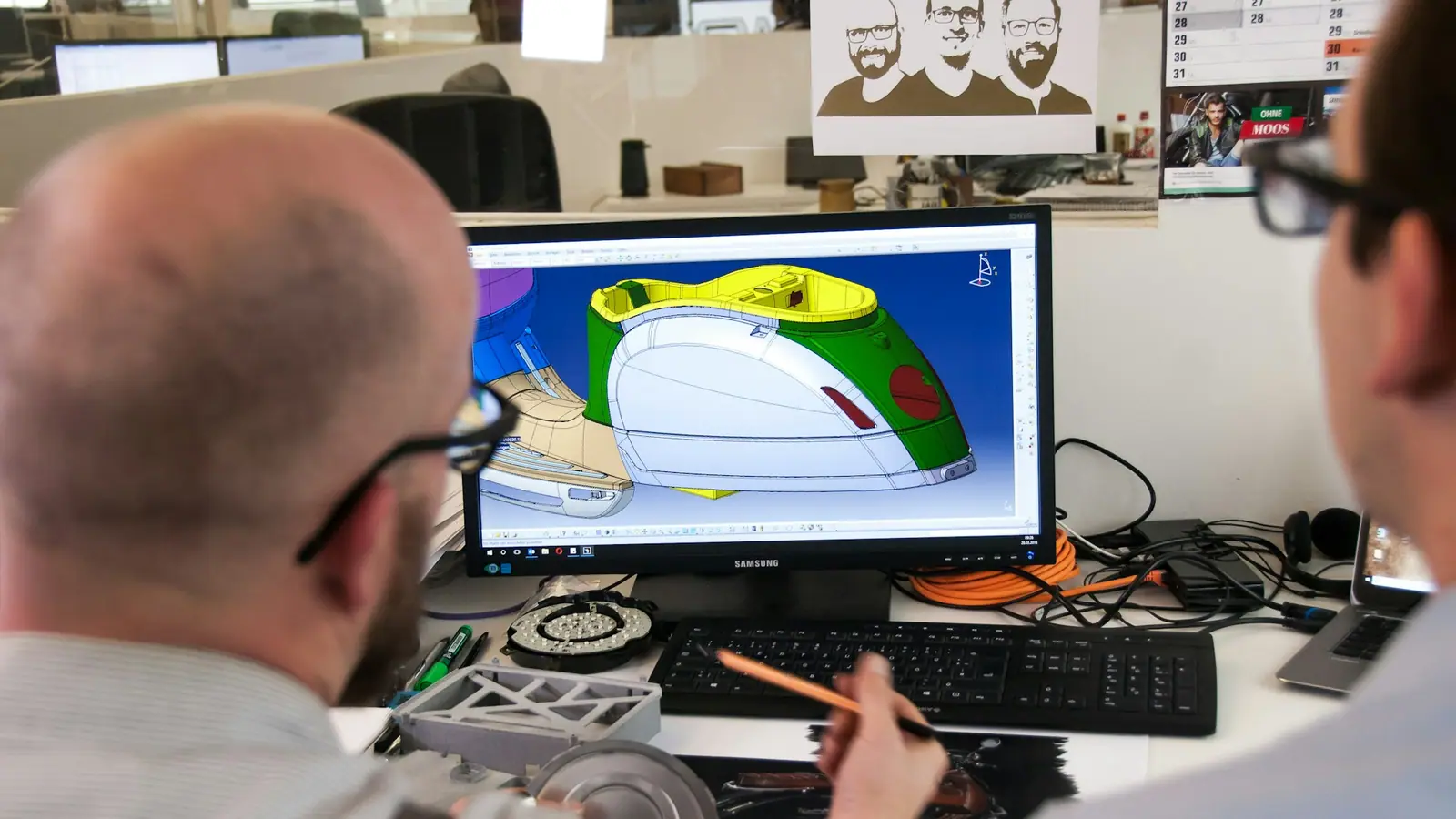 Two engineers reviewing a coloured CAD model of a moulded plastic part on a monitor.