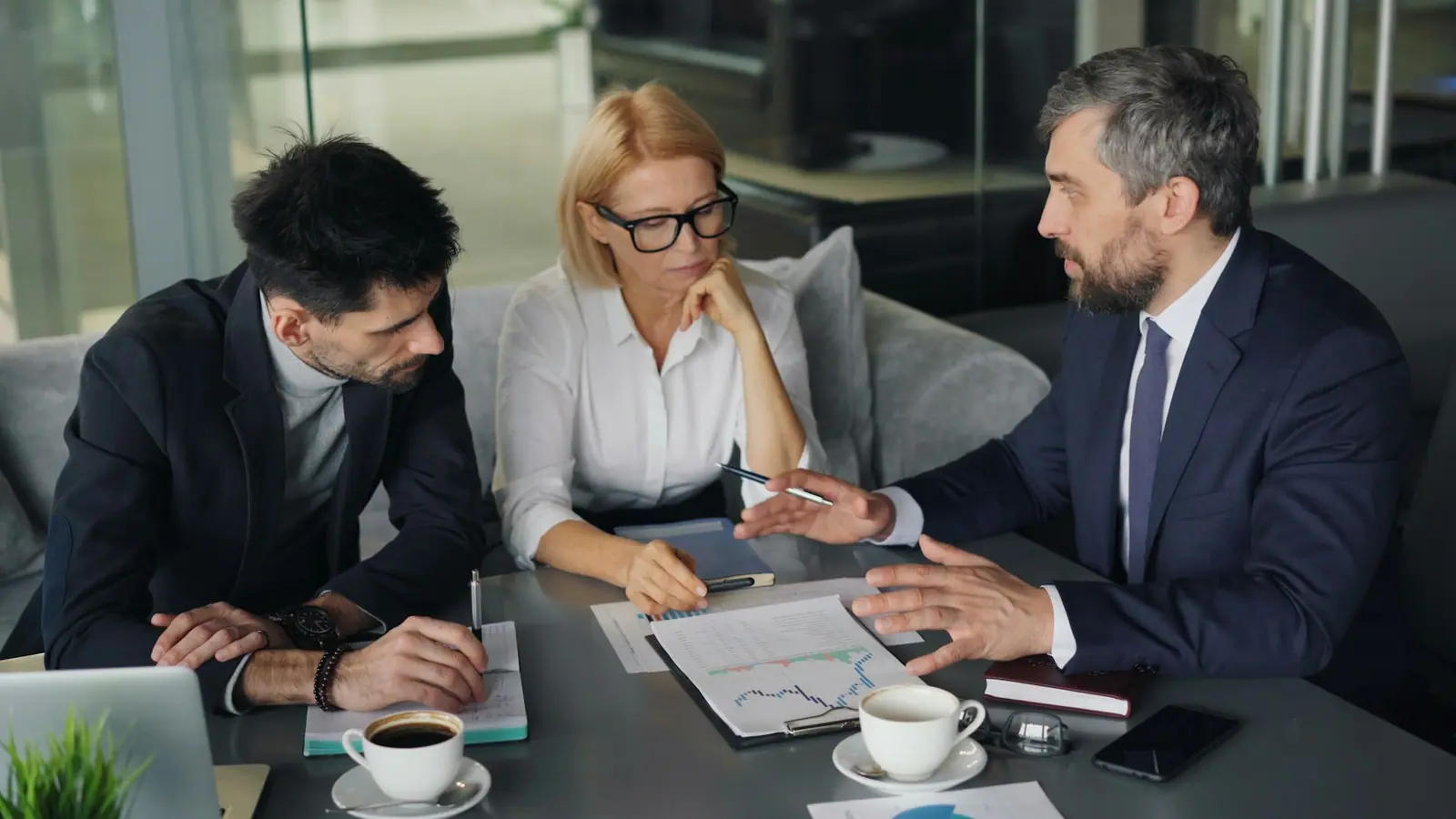 A buyer and two supplier representatives reviewing printed cost charts across a meeting table.
