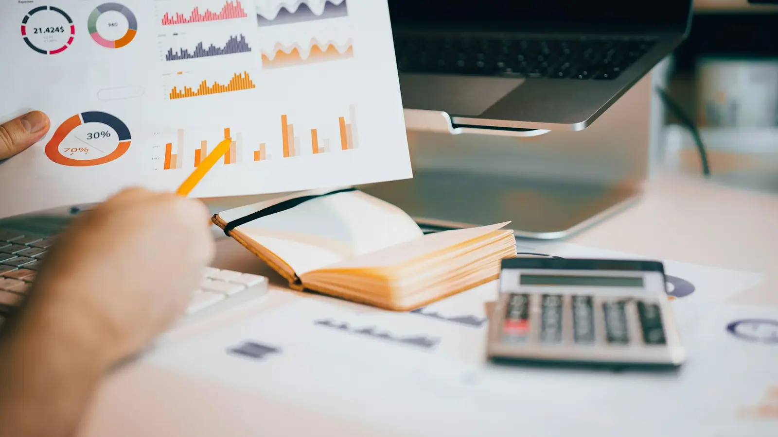 A hand holding a printed cost chart above a laptop, next to a calculator and an open notebook.