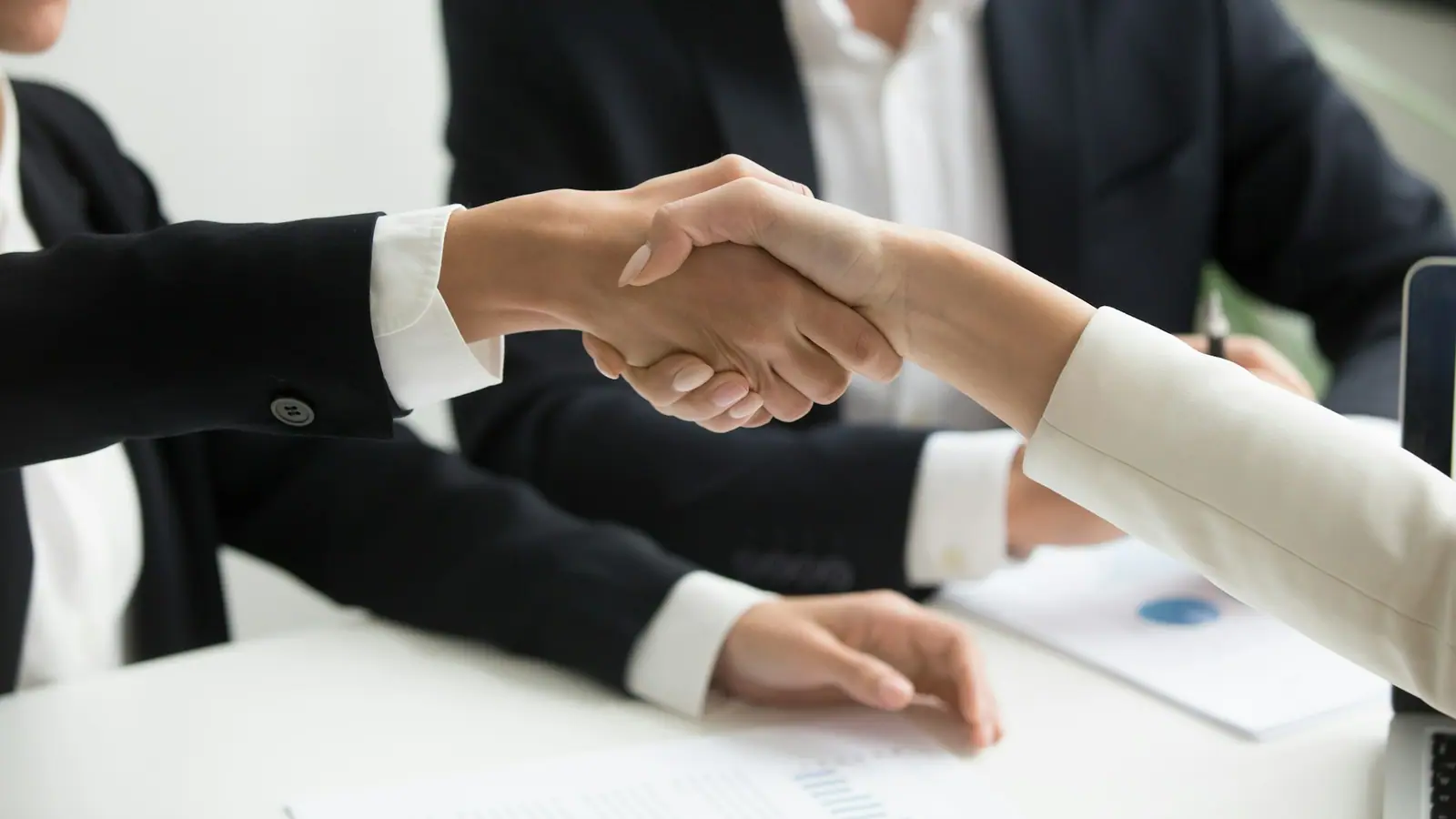Two people in business clothes shaking hands across a table.